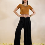 Woman wearing a mustard top and black pants with a gold necklace on a beige background